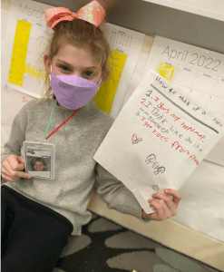 Student holding up her handmade teacher badge and anchor chart titled, "How to think of a topic."