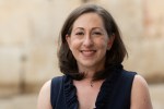 Stacey Shubitz is wearing a navy dress and is standing in front of a golden wall.
