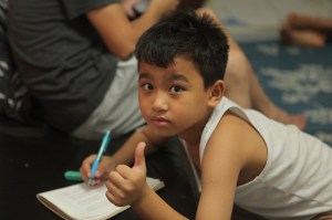 Boy in an undershit writing while lying on the floor with a pencil in one hand a thumbs-up in the other.