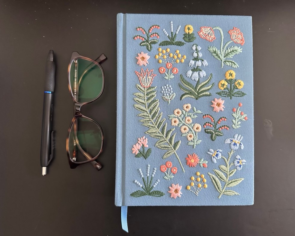 A blue floral notebook with a pen and reading glasses beside it.